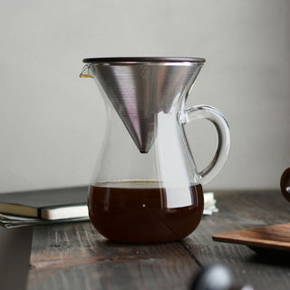 A clear glass pour-over coffee carafe with a stainless steel cone filter, filled with dark coffee, set on a wooden table.