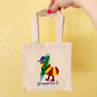 Hand holding mini canvas tote with colorful bug holding a pen against a yellow background