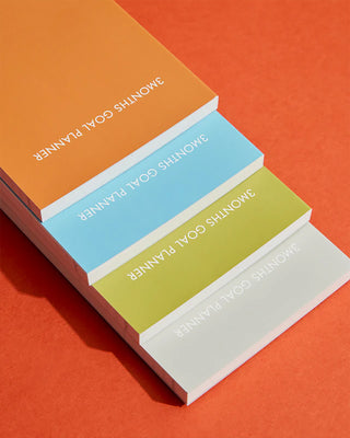 Stack of '3 Months Goal Planner' books in different colors on an orange background