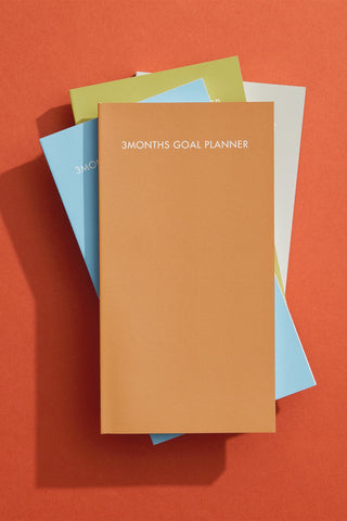 Stack of colorful planners with a focus on an orange one labeled '3 Months Goal Planner' on an orange background.