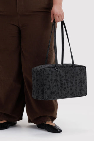 Medium nylon bowler bag with top handles, charcoal floral pattern, held at side.