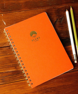 Orange notebook with the Beams Japan logo on a wooden surface, accompanied by two pencils.