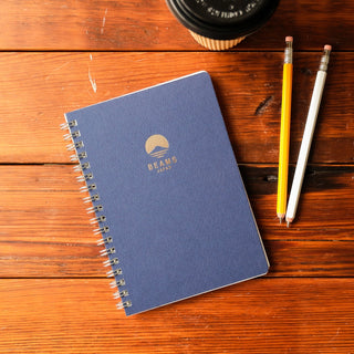 Blue notebook with the Beams Japan logo on a wooden surface with two pencils and a coffee cup.