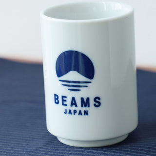 White ceramic mug with blue 'Beams Japan' logo on a blue textured surface