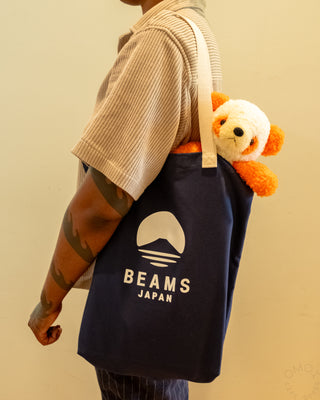 Person holding a navy tote bag with 'Beams Japan' logo and a plush toy, against a beige background