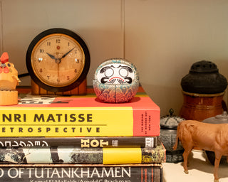 Stack of books with a clock and a green daruma surrounded by decorative items, against a neutral background