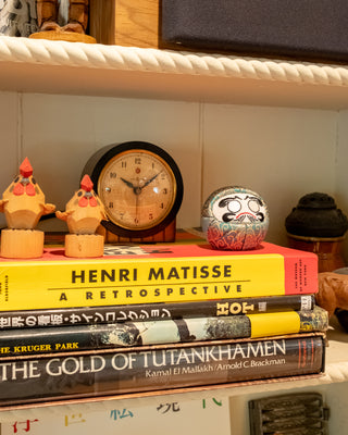 Stack of books on a shelf with decorative items including a clock a green daruma