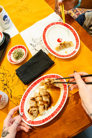 Two people enjoying dumplings with chopsticks on a table with an orange and white tablecloth.