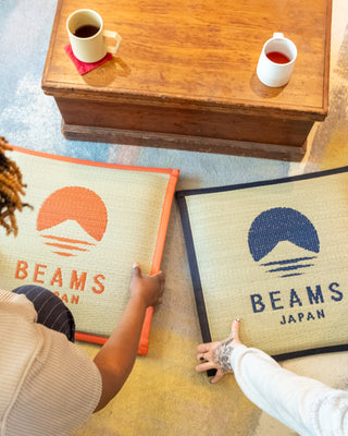 Two people holding BEAMS Japan mats on the floor with a wooden table in the background.