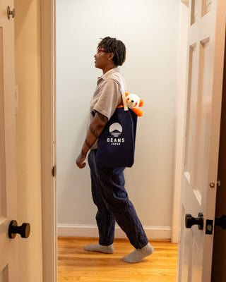 Person wearing a navy 'Beans Japan' tote bag with a stuffed bear, standing in a doorway.