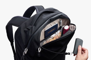 Black backpack with small compartment open showing travel items with a hand holding keychain on a white background