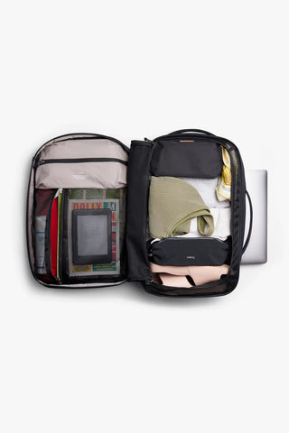 An open black backpack with travel items including a laptop, folded clothing, toiletry pouch, on a light gray background.