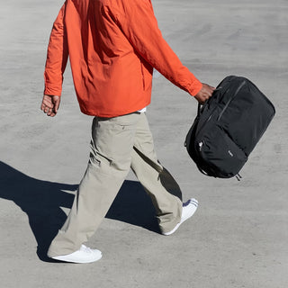 Person in orange jacket and beige pants walking with a black travel pack