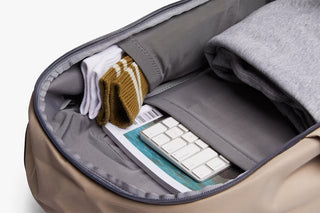 Close-up of an backpack showing its interior organization with a laptop sleeve holding a white keyboard and magazine, alongside folded clothing and socks.