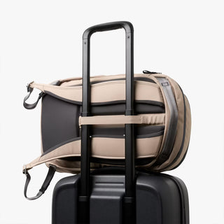 Beige backpack with black straps on top of a black suitcase on a white background