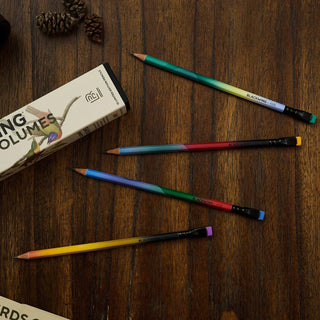 Four colorful pencils with a gradient design arranged on a dark wooden table next to a product box labeled 'Blackwing Volumes'.