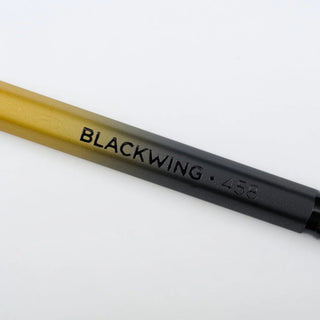 A close-up of a pencil with a gradient color transition from yellow to black, featuring the text 'BLACKWING 458'