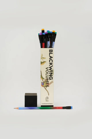 A set of colored pencils in a tall cream-colored box labeled 'BLACKWING VOLUMES', shown with colorful erasers and a single pencil laid out horizontally on a light gray background.
