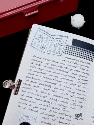 Open notebook with handwritten text and illustrations on a dark surface, with tool box and figurine in the background