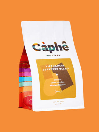 A bag of Câphé Roasters Vietnamese Espresso Blend coffee on a solid orange background. The packaging is primarily white with a large yellow label on the front and a colorful striped pattern visible on the left side.