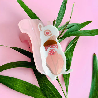 Squirrel shaped hairclip holding an acorn against a plant on a pink background