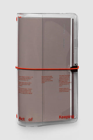 A clear plastic journal cover holding a taupe-brown paper journal, secured with a bright orange elastic band. The journal features minimalist orange/red text, and the cover itself has orange text reading 'Act of Keeping'.