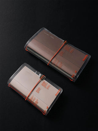 Two clear plastic notebook covers with orange elastic bands, holding brown and beige notebooks, displayed on a dark background.