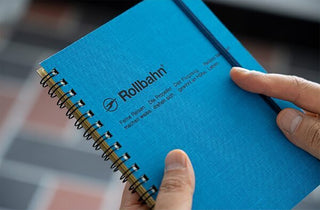 Blue spiral notebook close-up with Rollbahn logo and hands holding the cover