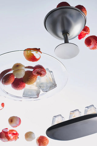 Fruit in a glass bowl with jelly cubes, surrounded by lychees on a white background.