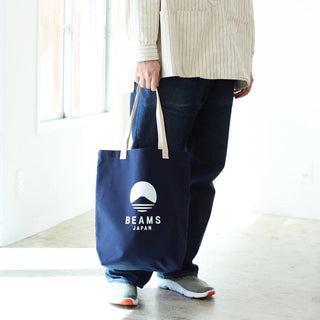 Person holding a navy tote bag with 'BEAMS JAPAN' logo in a minimal indoor setting.