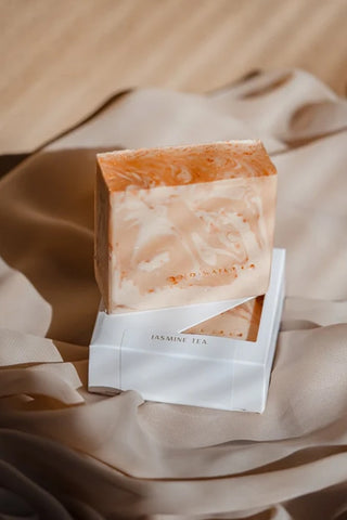 A rectangular bar of marbled soap in peach, cream, and orange tones, displayed on top of a white box labeled 'JASMINE TEA' against draped beige fabric.