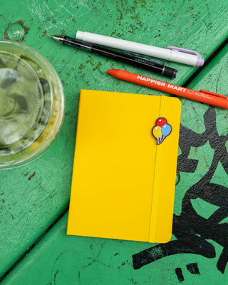 A bright yellow planner with an elastic closure and a small colorful balloon design, styled on a green wooden surface with pens and a drink nearby.