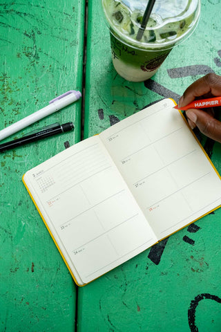 An open weekly planner notebook with cream pages on a green wooden table. A hand holds an orange pen above it, with an iced matcha, a white pen, and a black mechanical pencil nearby