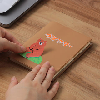 A person holding a brown notebook on a wooden desk. The notebook features a cartoon bear sticker and Japanese text, with a laptop partially visible in the top left corner.