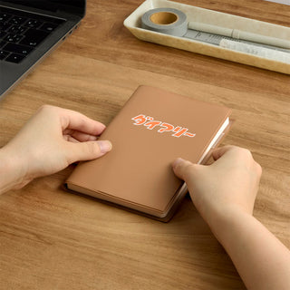 A person's hands holding a brown planner with orange and white Japanese text on the cover, placed on a wooden desk next to a laptop and stationery tray.
