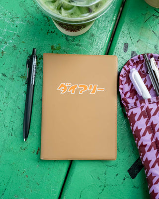A brown planner cover with Japanese text in orange and white on the cover, styled on a green wooden table next to a black pen and a pink patterned case.