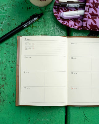 An open weekly planner on a green wooden surface with pens and a patterned pouch nearby.