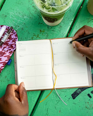 A top-down view of a person writing in an open weekly planner with a black pen, set on a green wooden table next to a green drink and a patterned pouch.