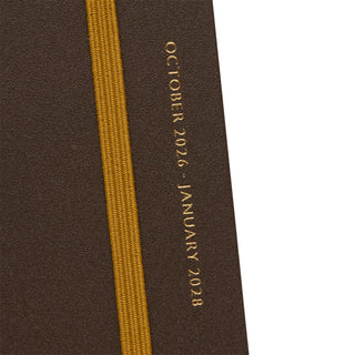 Close-up of a dark brown textured planner cover with a vertical orange elastic band and gold text reading 'OCTOBER 2026 - JANUARY 2028' on a white background.