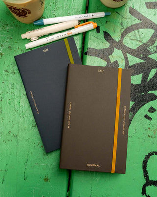 Planner covers with elastic closures on a green wooden table with pens and coffee cups visible. One planner is dark brown with an orange band; the back planner is dark blue with a green elastic band.