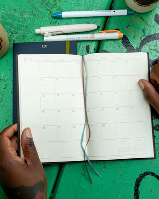 An open monthly planner with blank calendar pages is held by two hands over a green wooden table. Several pens are placed above the planner.