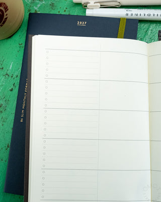 An open monthly planner showing blank lined pages with checkboxes, placed on a green wooden table with part of a pen and iced coffee cup visible.