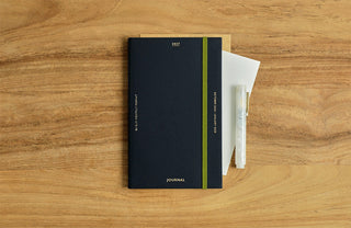 A navy blue planner with a green elastic band closure, shown on a wooden tabletop alongside a white pen and a folded white paper.