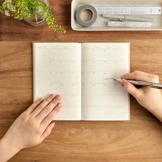 An open monthly calendar planner on a wooden desk, with a person writing. A tray with small stationery items and a small plant is visible in the background.