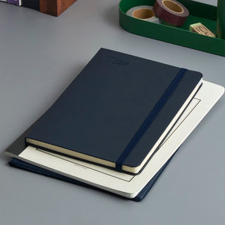 A dark hardcover notebook with an elastic closure strap, stacked on top of a white notebook with stationery items in the background of the desk