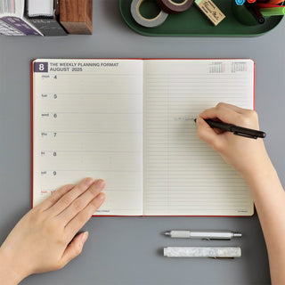 An open weekly planner with lined pages on a gray desk, with a person's hands writing with a pen