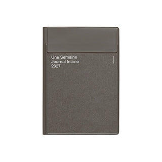 A dark gray planner with a matching flap closure on the cover, featuring white text that reads 'Une Semaine Journal Intime 2027' on a plain white background.