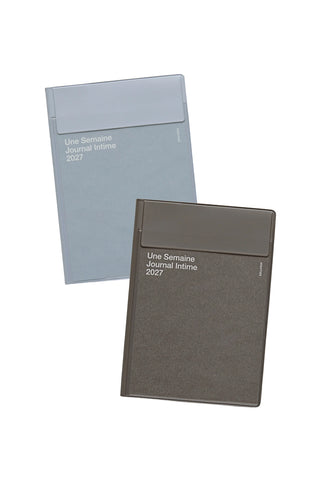 Two planners shown on a plain white background, one in pale blue-gray and one in dark gray. Both have a pocket cover and white printed text reading 'Une Semaine Journal Intime 2027'.