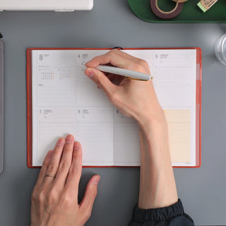 A top-down view of a person writing in an open weekly planner shown on a gray desk with other desk accessories partially visible.