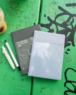 Two copies of the 'Une Semaine Intérieure 2027' journal, one in dark gray and one in light gray, displayed on a green wooden surface. White pens are placed next to them, and a partial iced coffee cup is visible in the top-left corner.